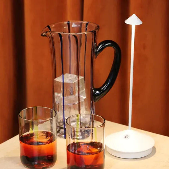 Glass pitcher and cups with a white lamp on a wooden surface against an orange curtain.