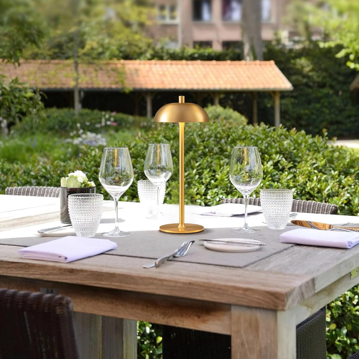 Outdoor dining setup with a wooden table, gold lamp, and glasses in a garden setting.