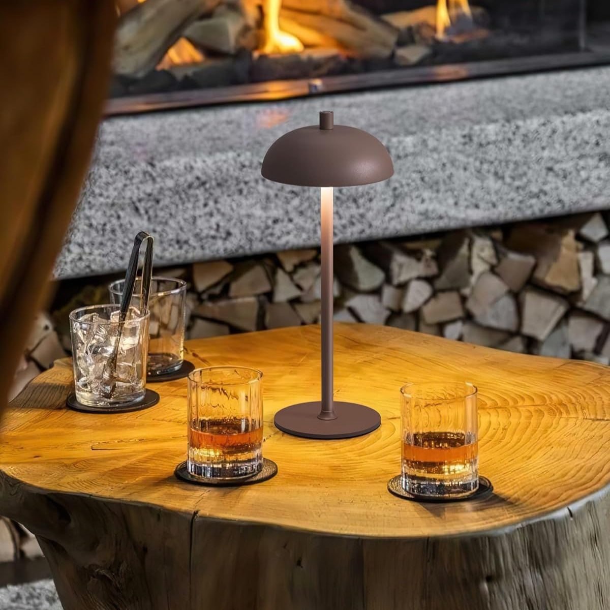 Table lamp on a wooden table with glasses of whiskey in front of a fireplace.