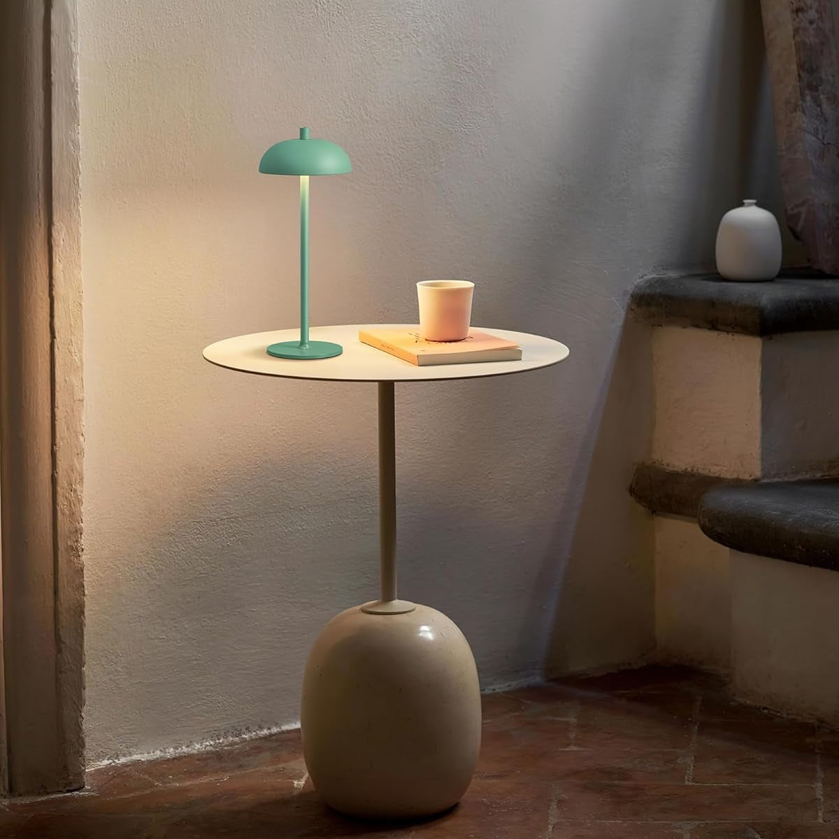Small round table with a green lamp and a pink cup against a neutral wall.