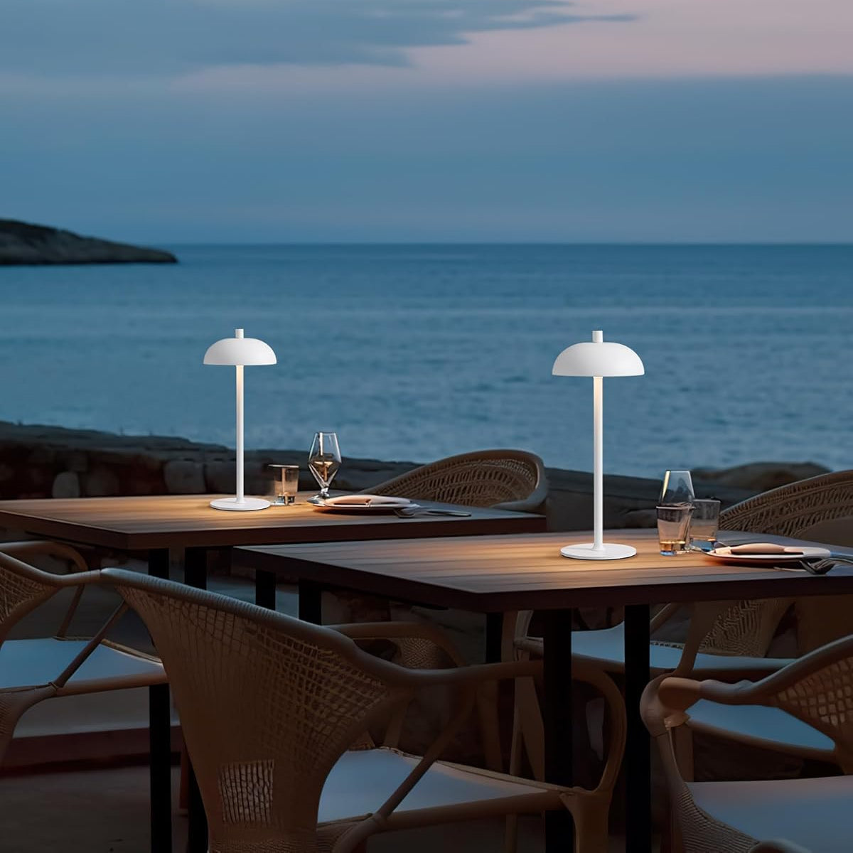 Outdoor dining setup with two lamps on a table by the sea at dusk.