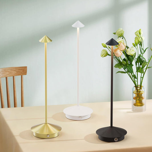 Three table lamps in gold, white, and black on a wooden table with flowers in the background.