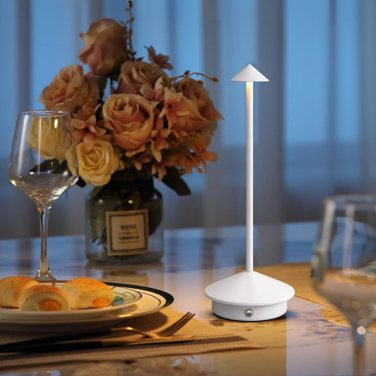 White LED table lamp on a dining table with a glass of wine, plate of food, and vase of flowers.