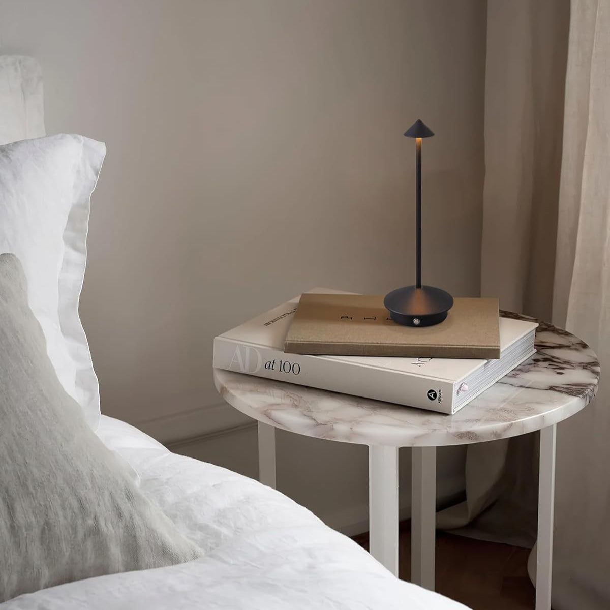 Small round marble table with a lamp and books next to a bed in a bedroom.